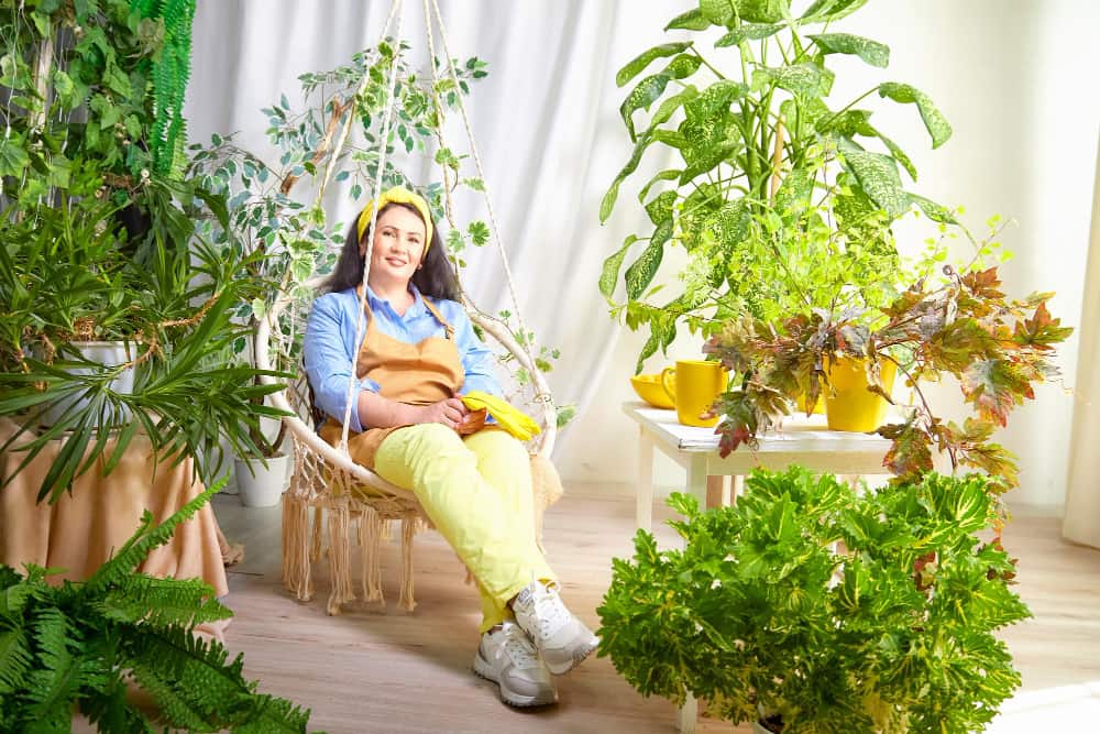 Une femme relaxée assise dans un fauteuil suspendu en macramé au milieu d'un salon de jardin intérieur luxuriant pour aménager un jardin bien-être