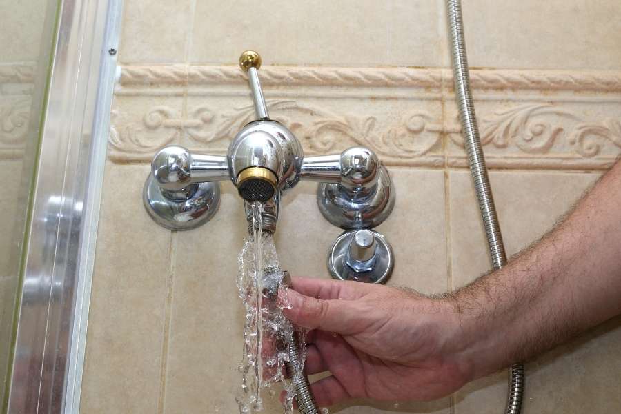 Une main visse ou dévisse le raccord d'un flexible de douche directement sous un robinet mélangeur mural pendant que l'eau s'écoule.